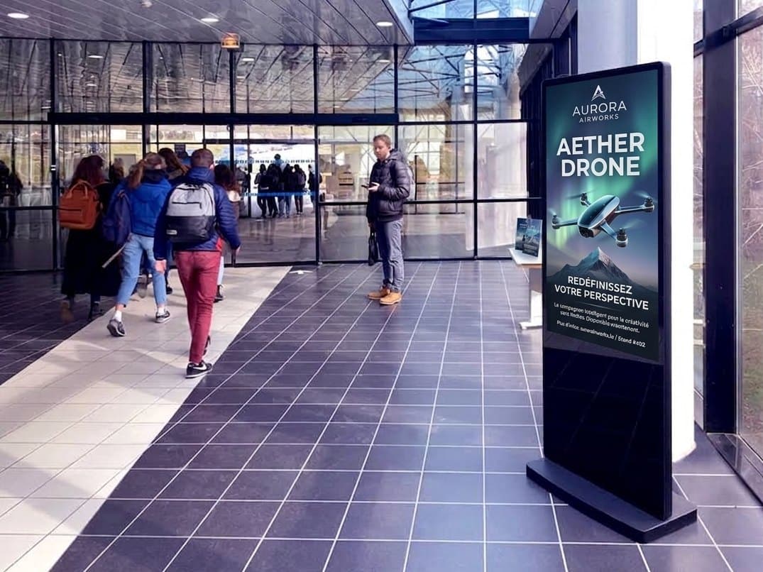 Un totem publicitaire dans un hall, devant lequel passent des visiteurs